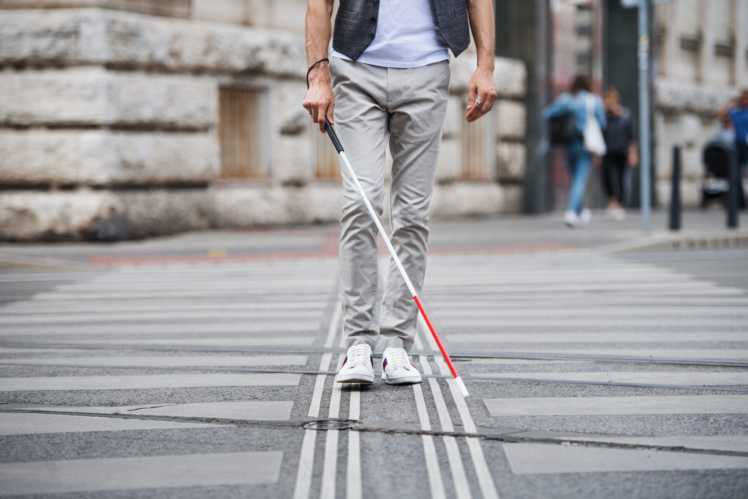 Zjęcie przedstawia osobę niewidomą z laską od pasa w dół, przechodzącą przez przejście dla pieszych.
