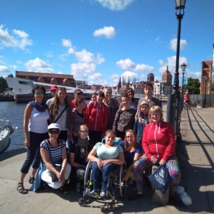 Grupa około piętnastu młodych osób, pozuje na deptaku w Gdański. Wśród nich w pierwszym rzędzie jest młoda kobieta na wózku. W tle widać rzekę i statki oraz starówkę miasta.