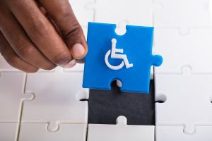 Niebieski puzzel z symbolem osoby z niepełnosprawnością trzymany w ręce na tle białych puzzli.