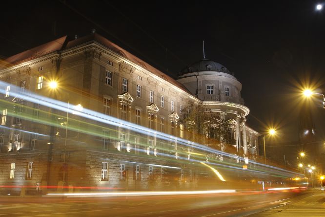 Zdjęcie przedstawia budynek Collegium Maius nocą. Przed budynkiem jadący tramwaj z efektem ruchu w przestrzeni.