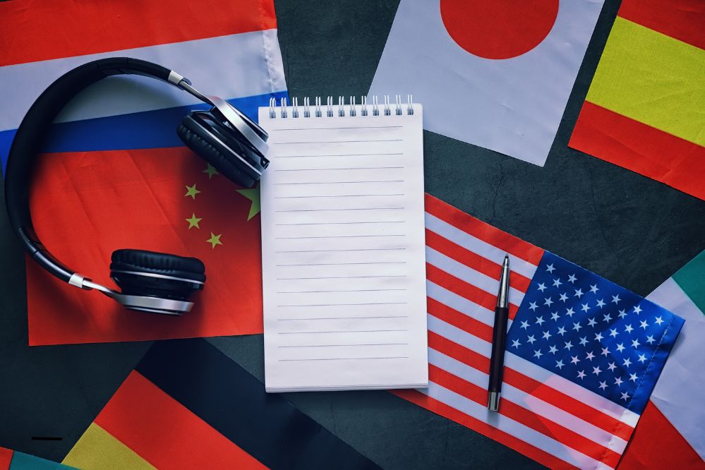 Rozłożone na stole flagi Japonii, Hiszpanii, USA, Niemiec, na nich leżą słuchawki, notes oraz długopis.
