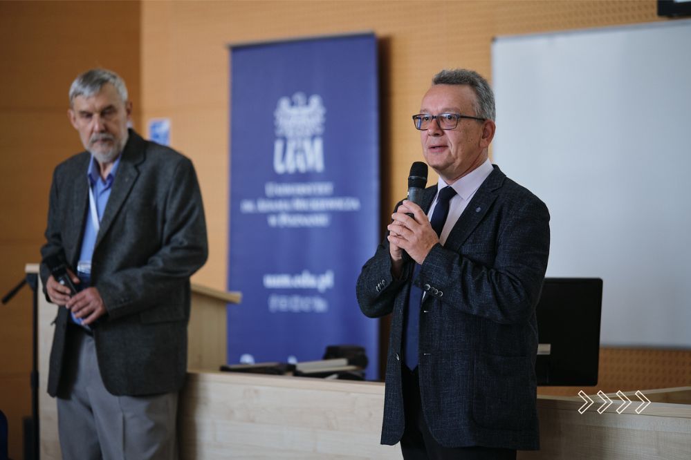 Dwóch mężczyzn stoi przed katedrą na sali wykładowej. Jeden z nich przemawia.